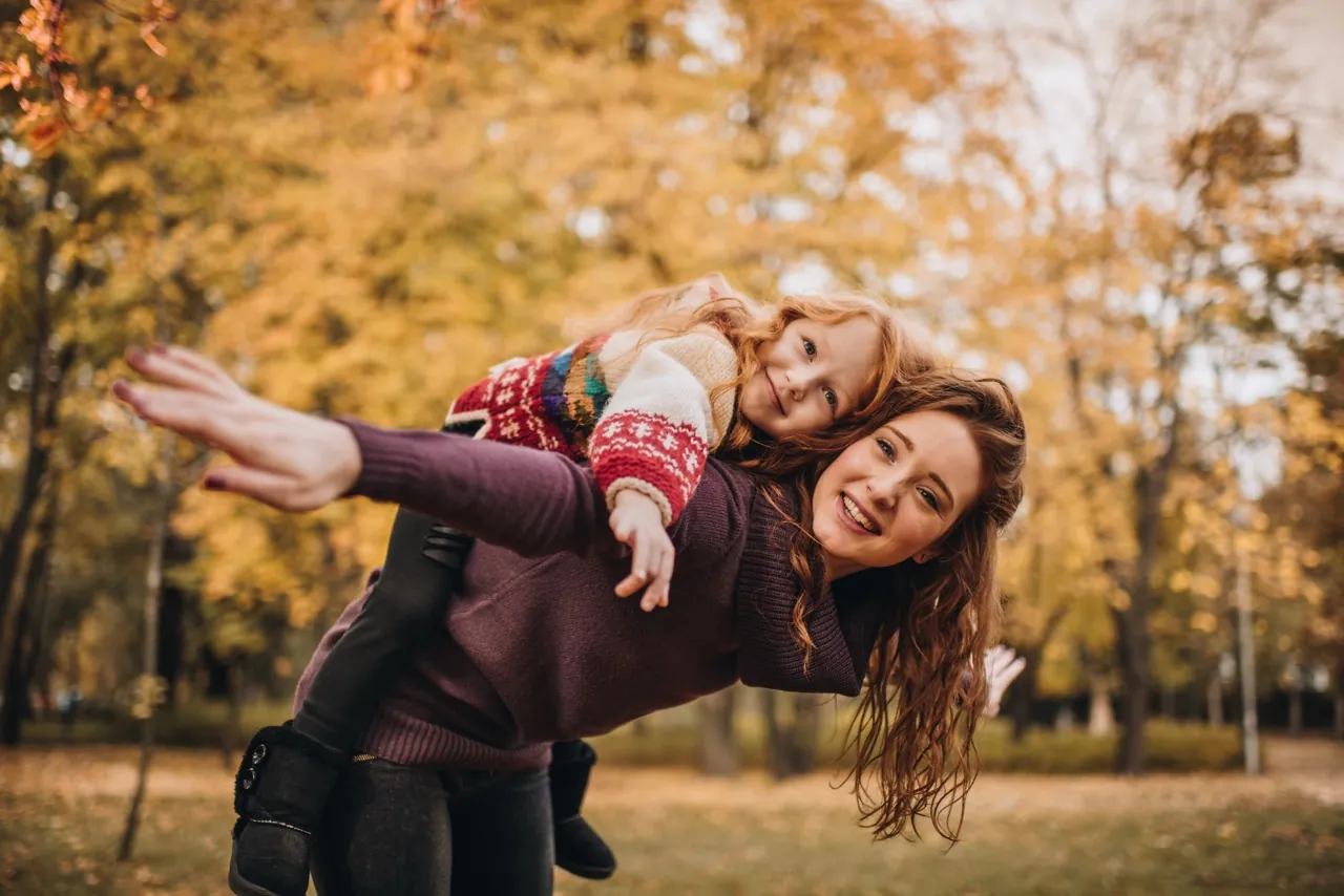 Babysitterin trägt ein lachendes Mädchen huckepack durch den Herbstpark