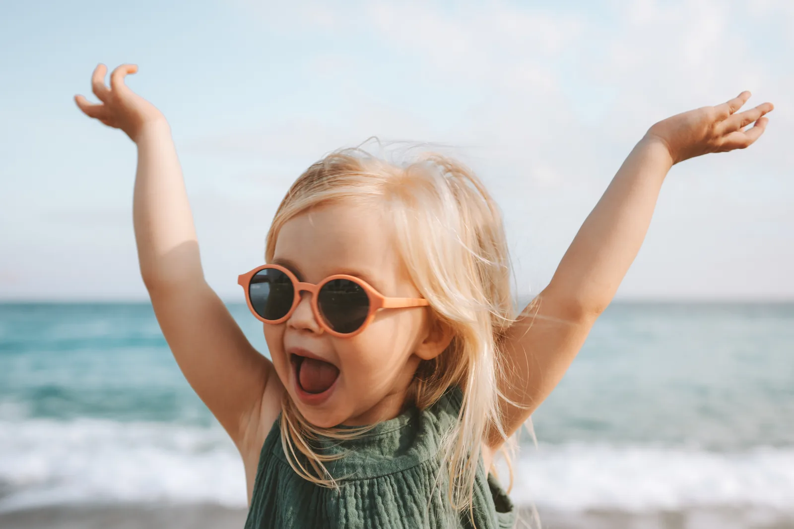 Lustiges kleines Mädchen spielt draußen und überrascht emotionales Kind mit Sonnenbrille, 3 Jahre altes Baby hebt die Hände, Familienurlaub