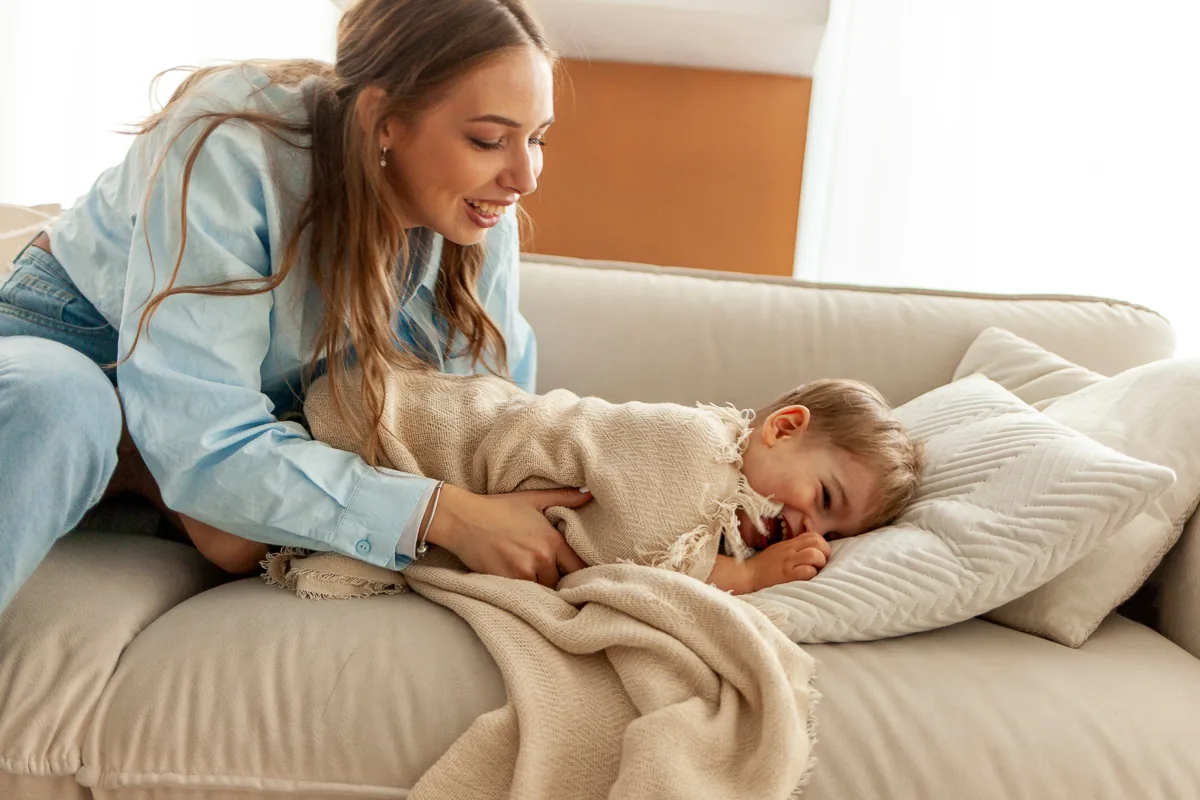 Nanny spielt mit einem Kind zu Hause auf dem Sofa und deckt ihn mit einer Decke zu, der 2-jährige Junge lacht und hat Spaß